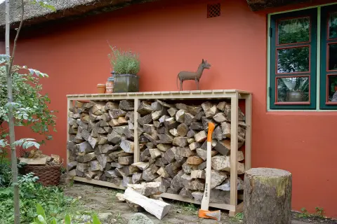 Praktische brennholz lagern ideen: Holzstapel in einem Holzregal vor einer roten Wand, daneben ein Baumstumpf und eine Axt.
