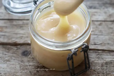 Un cucchiaio preleva il cremoso latte condensato da un barattolo di vetro su un tavolo di legno rustico.