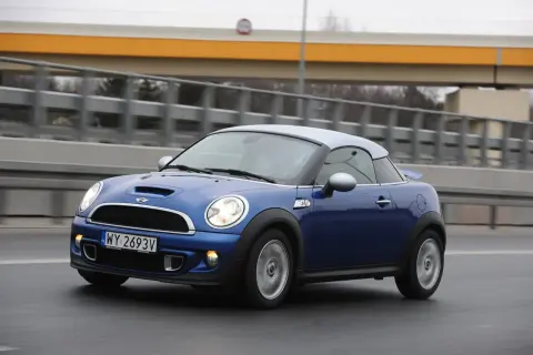 Niebieski Mini R58 Cooper S z szarym dachem pędzi po drodze, za nim widać most.