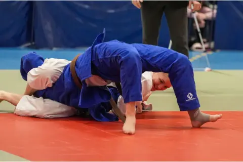 Två judokas i blå och vita dräkter kämpar på en röd matta. En av dem, med ett leende, utför en rörelse inspirerad av Jigoro Kano.