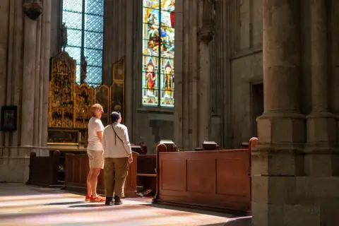 Zwei Besucher bestaunen die bunten Glasfenster im Kölner Dom. Sonnenlicht fällt durch die Fenster und malt bunte Muster auf den Boden.