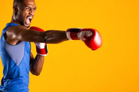 A boxer in a blue tank top throws a punch, his face contorted in a roar. He's ready to compete in the junior welterweight weight class.
