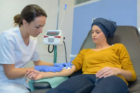 Eine Krankenschwester bereitet eine Infusion vor. Die Patientin, die eine Kopfbedeckung trägt, erhält die Behandlung, ein Zeichen dafür, dass die Chemo wirkt.