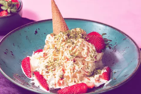Spaghetti-Eis mit Erdbeeren und Pistazien in einer blauen Schale, garniert mit einer Eistüte.