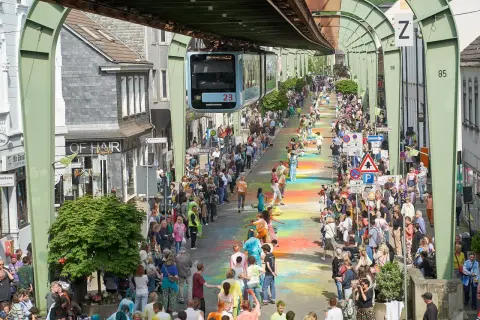Bunte Farben auf dem Boden und eine Schwebebahn über der belebten Straße im Wuppertal Zentrum. Menschenmassen säumen die bunte Gasse.
