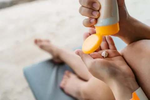 Manos aplicando crema solar caduca en la palma. Un niño descansa en una tumbona al fondo.
