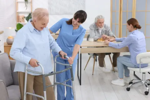 Une aide-soignante accompagne une résidente d'un EHPAD Alzheimer avec un déambulateur. Au fond, d'autres résidents sont à table.