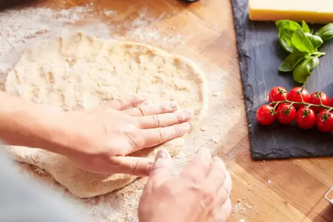 Hände formen den **pizzateig auftauen** auf einer bemehlten Holzoberfläche. Daneben liegen frische Tomaten, Mozzarella und Basilikum.