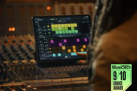A person's finger touches a tablet displaying Logic Pro for iPad, showing a colorful grid of musical patterns, in a recording studio.