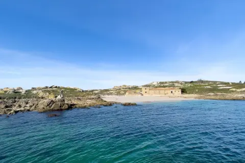 Vista de la isla de Sálvora con sus rocas, una playa de arena y un edificio de piedra bajo un cielo azul.
