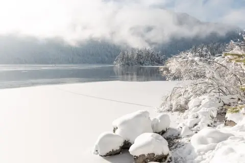 Eibsee Winter: Eisbedeckter See, schneebedeckte Bäume und Felsen unter nebligen Bergen. Ein friedlicher Anblick.