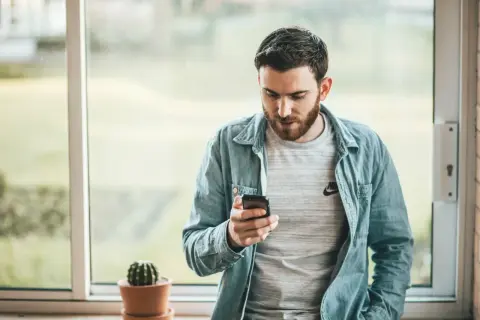 Hombre con barba mira su móvil, quizás buscando frases cuando no te responden. Un cactus en maceta decora el alféizar.