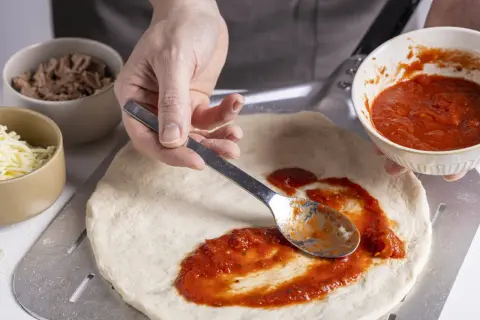 Zubereitung eines italienischen Thunfisch Pizza Rezepts: Tomatensauce wird auf den Teig gestrichen. Daneben liegen Thunfisch und Käse.