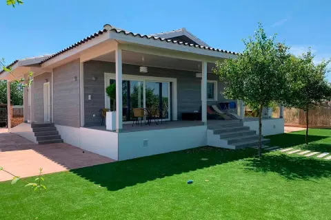 Moderna casa Canexel con porche, césped artificial y árboles frondosos bajo un cielo azul.