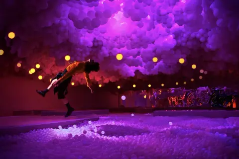 A person in a yellow shirt leaps into a ball pit at the balloon museum London, surrounded by purple clouds and yellow lights.