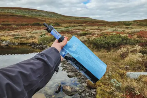 Vattenrening vandring: en hand håller en Sawyer vattenfilterpåse vid en bäck i fjällterräng.