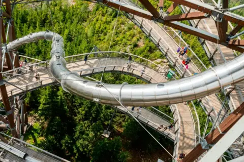 Drewniana ścieżka w koronach drzew z metalową zjeżdżalnią, widok z góry na Sky Walk w Czechach.