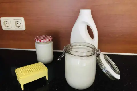 Zelf wasmiddel maken met dille en kamille. Glazen potten met wit wasmiddel, een gele rasp en een witte fles staan op het aanrecht.