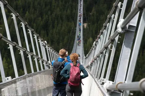 Para idzie przez wiszący most w Czechach, robiąc zdjęcia. W tle widać las i innych turystów.