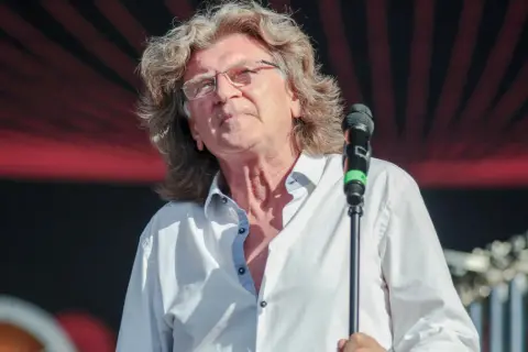 Zbigniew Wodecki śpiewa "Lubię wracać tam, gdzie byłem", z mikrofonem w dłoni, na tle czerwono-czarnej sceny.