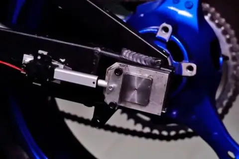 Mecanismo de ajuste para cadena destensada moto, con actuador lineal y soporte metálico sobre basculante azul.