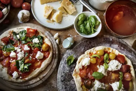 Dwie pizze z sosem pomidorowym, świeżymi pomidorkami, bazylią i kremową włoską mozzarellą. Obok składniki: parmezan, czosnek, pomidory.