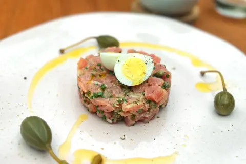 Un délicieux tartare de thon, garni d'un œuf de caille et de câpres, est servi sur une assiette blanche avec un filet d'huile d'olive.