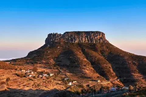 La imponente fortaleza de Chipude se alza sobre un paisaje rural con casas blancas y palmeras, bajo un cielo azul claro.