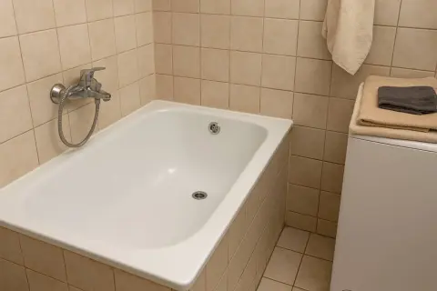 Biała wanna w łazience z beżowymi płytkami. Czy renowacja wanny to dobry pomysł? Obok pralka z ręcznikami.