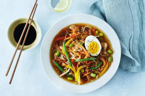 En skål med nudlar, grönsaker, kyckling och ett halvt ägg. Soppan innehåller morötter, sockerärtor, böngroddar och paprika.