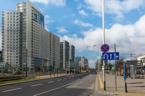 Ulica z nowoczesnymi budynkami i tramwajem. Na słupie widoczny znak D3 informujący o zakazie zatrzymywania się i dozwolonych kierunkach jazdy.