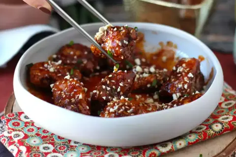 Chrupiący kurczak gochujang w sosie, posypany sezamem i orzeszkami, podany w białej misce.