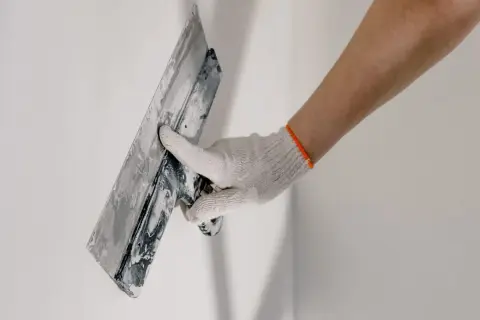 Mano enguantada aplicando material para revocar pared con llana.