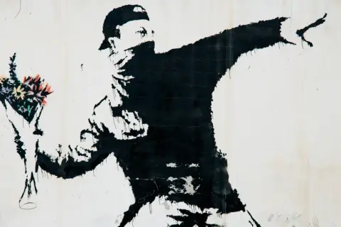Czarny mural przedstawia zamaskowanego mężczyznę w czapce, rzucającego bukiet kolorowych kwiatów. To najładniejszy rysunek na świecie, pełen nadziei i buntu.