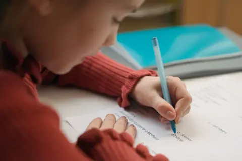 Dziecko w czerwonym swetrze pisze długopisem po kartce. Czy to zadanie z języka angielskiego, od jakiej średniej jest 5?