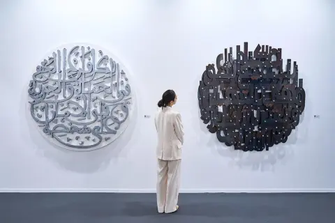 En kvinna i ljus kostym betraktar två runda konstverk med arabisk kalligrafi. Det vänstra är i metall, det högra i mörkare material. En atra daniel.