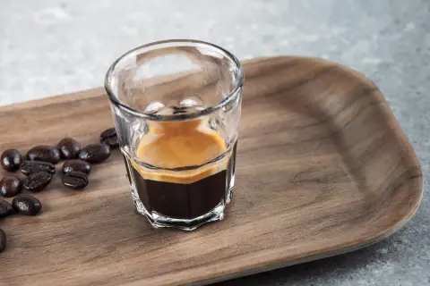 En liten shot med espresso, med crema, bredvid kaffebönor på en träbricka. En perfekt start på dagen, en stark espresso vs. vanligt kaffe.