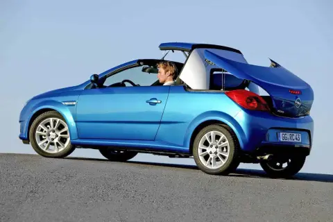 Niebieski kabriolet Opel Tigra z otwartym dachem, kierowca w środku, na tle błękitnego nieba.