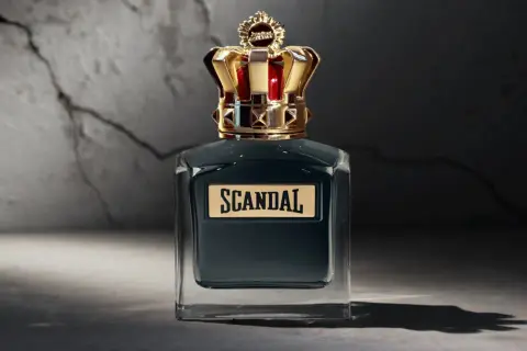 Elegancka butelka perfum Scandal męskie z koroną na nakrętce, stojąca na tle pękniętej ściany.