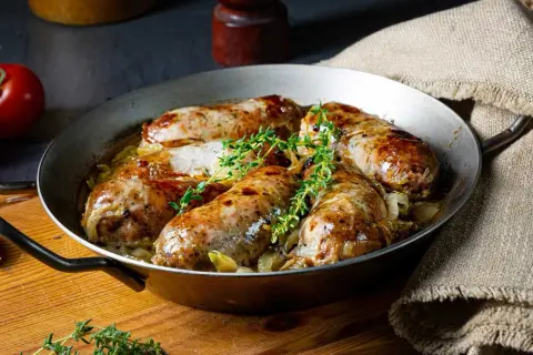 Pieczona w piekarniku biała kiełbasa surowa z cebulą i tymiankiem, podana w metalowej patelni.