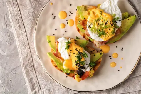 Två läckra eggs benedict på rostat bröd med avokado, bacon och hollandaisesås. Garnerade med persilja och svarta sesamfrön.
