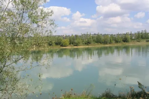 Le lac de Beaupuy, paisible sous un ciel bleu parsemé de nuages blancs, reflète la végétation luxuriante de ses rives.