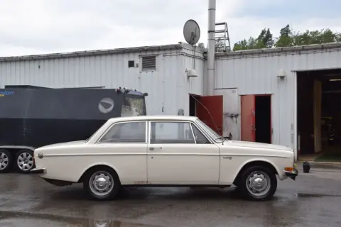 En vit Volvo 142 från 1973 står parkerad framför en industribyggnad.
