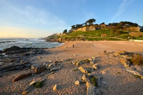 Atardecer en la playa concheira de Baiona. Olas rompen en la costa rocosa, mientras una fortaleza histórica corona la colina verde.