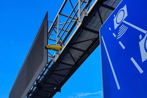Żółta kamera na konstrukcji nad drogą, obok niebieskiego znaku z symbolami aparatów. To zapowiedź nowych odcinkowych pomiarów prędkości.