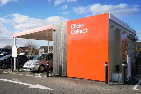 Zona de **click and collect** con coches aparcados bajo un toldo. Edificio naranja brillante con el texto "Click+ Collect".