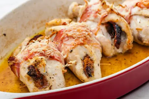 Pyszne roladki z kurczaka zawinięte w boczek, zapieczone z suszonymi pomidorami i posypane parmezanem, w sosie własnym.