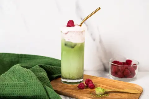 Orzeźwiające matcha z malinami, ozdobione pianką i świeżymi malinami, z dodatkiem proszku matcha na desce.