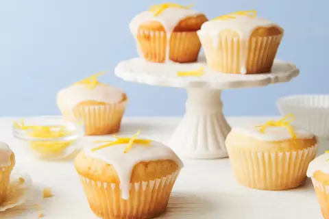 Saftige vegane Zitronenmuffins mit Zuckerguss und Zitronenschale verziert, auf einem weißen Kuchenständer und Tisch.