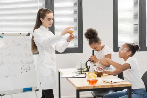 Nauczycielka demonstruje doświadczenie chemiczne, podczas gdy uczniowie pracują z mikroskopem i probówkami. Na tablicy widać wzory chemiczne, pomocne przy konkursy przedmiotowe arkusze.